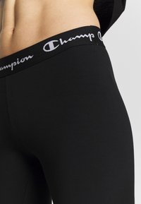 Czarne legginsy wykonane z gładkiego, elastycznego materiału z szerokim czarnym pasem w talii ozdobionym białym napisem "Champion".
