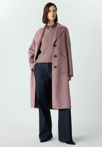 Długi, pastelowo-różowy wełniany płaszcz z klapami, dwoma przednimi kieszeniami i czarnymi guzikami, noszony na beżowym, dzianym swetrze i ciemnoniebieskich rozszerzanych spodniach.
