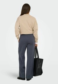 Beige sweatshirt med faldne skuldre, parret med mørkegrå brede bukser. Sort læder tote taske holdt i venstre hånd, flade sko synlige.