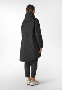 Sort quilted hættetrøje med en lang silhuet, der har et tekstureret mønster, sidelommer og en buet kant, parret med mørke bukser og sneakers.