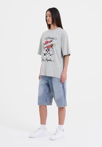 Camiseta gris de gran tamaño con estampado gráfico que presenta un diseño de calavera y corazón, combinada con pantalones cortos de mezclilla azul claro y zapatillas blancas.