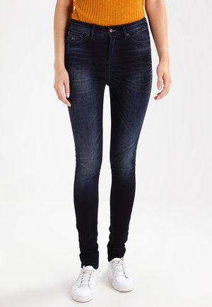 Femme portant un jean skinny bleu foncé, un haut sans manches côtelé orange et des baskets blanches, debout devant un fond uni.