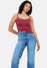 Kobieta z kręconymi włosami, ubrana w bezrękawnik w ciemnoróżowy kolor z koronkowym wykończeniem oraz w wysokie niebieskie dżinsy, pozuje z jedną ręką na biodrze.