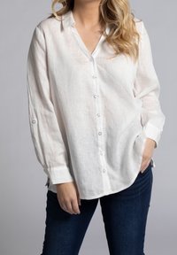 Camicia bianca di lino con scollo a V, chiusura con bottoni e maniche lunghe. Presenta una leggera texture e una vestibilità morbida, abbinata a jeans in denim scuri.