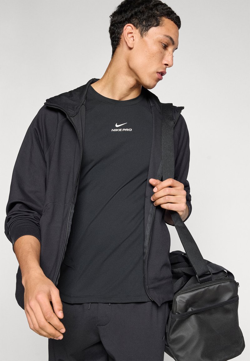 Black athletic outfit consisting of a zip-up hoodie and fitted shirt, both with Nike branding. A black satchel is held in the subject's hand.