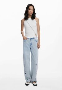 Witte mouwloze top met zijkant strikdetail, gecombineerd met lichtblauwe jeans met wijde pijpen en knoopaccenten langs de pijpen en voorzakken.