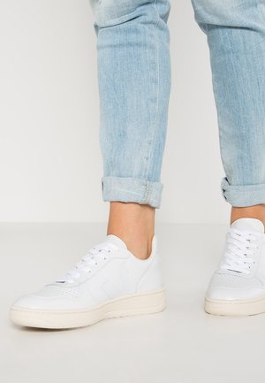 Pieds portant un jean bleu clair retroussé et des baskets blanches basses, debout sur une surface claire devant un fond uni.