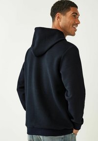 Marineblauwe, gestructureerde hoodie met een capuchon en een ruitpatroon. Geribbelde manchetten en zoom, gedragen door een persoon van achteren, met minimale zichtbare details.
