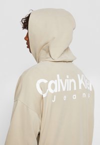 Béžová mikina s měkkou texturou, která má velké bílé logo "Calvin Klein Jeans" na zádech a stahovací kapuci.