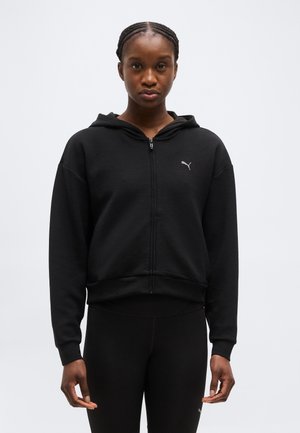 Woman wearing a black zip-up hoodie with a Puma logo on the chest and black leggings, standing against a plain light background.