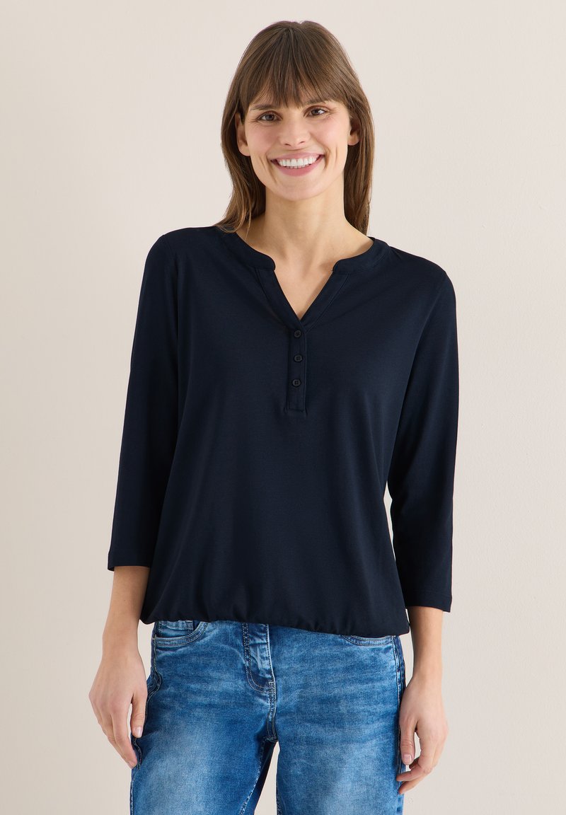 Femme souriante portant un t-shirt henley à manches longues bleu marine et un jean bleu, debout devant un fond clair uni.