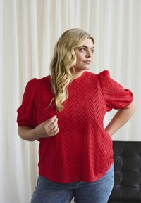 Femme blonde portant un chemisier rouge à manches courtes texturé et un jean bleu, se tenant avec une main sur la hanche et tenant une mèche de cheveux.