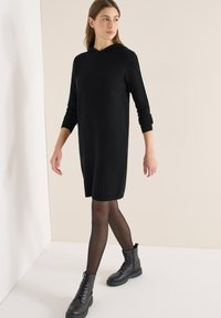 Vestito nero con cappuccio in maglia, maniche lunghe e spacchi laterali, abbinato a stivaletti neri con lacci e collant neri trasparenti.