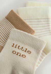 Tres pares de calcetines para bebé en tonos beige y crema; un par tiene "pequeñín" tejido en letras marrones en el puño.