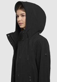 Veste à capuche noire en matériau lisse et résistant à l'eau, dotée d'une fermeture éclair et de boutons-pression, de cordons de serrage ajustables et d'un écusson logo.