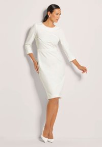 White dress with three-quarter sleeves, fitted shape, smooth texture, asymmetrical design on the front, paired with white pointed heels.