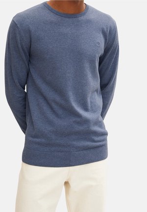 Homme portant un pull bleu à manches longues et col rond associé à un pantalon écru sur un fond uni.