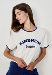 Joven mujer con cabello largo y castaño que lleva una camisa blanca con ribetes azules que dice "AMABILIDAD en el interior", ajustándose la camisa y mirando hacia abajo.