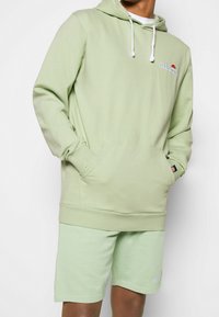 Felpa verde chiaro con coulisse, tasca e logo. Abbinata a pantaloni corti verde chiaro coordinati. Tessuto morbido e design casual.
