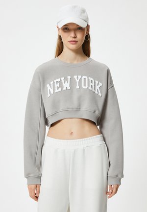 Grauer, kurzgeschnittener Sweatshirt mit "NEW YORK" in weißen Buchstaben, langen Ärmeln und einer strukturierten Oberfläche; kombiniert mit weißen Jogginghosen mit elastischem Bund.