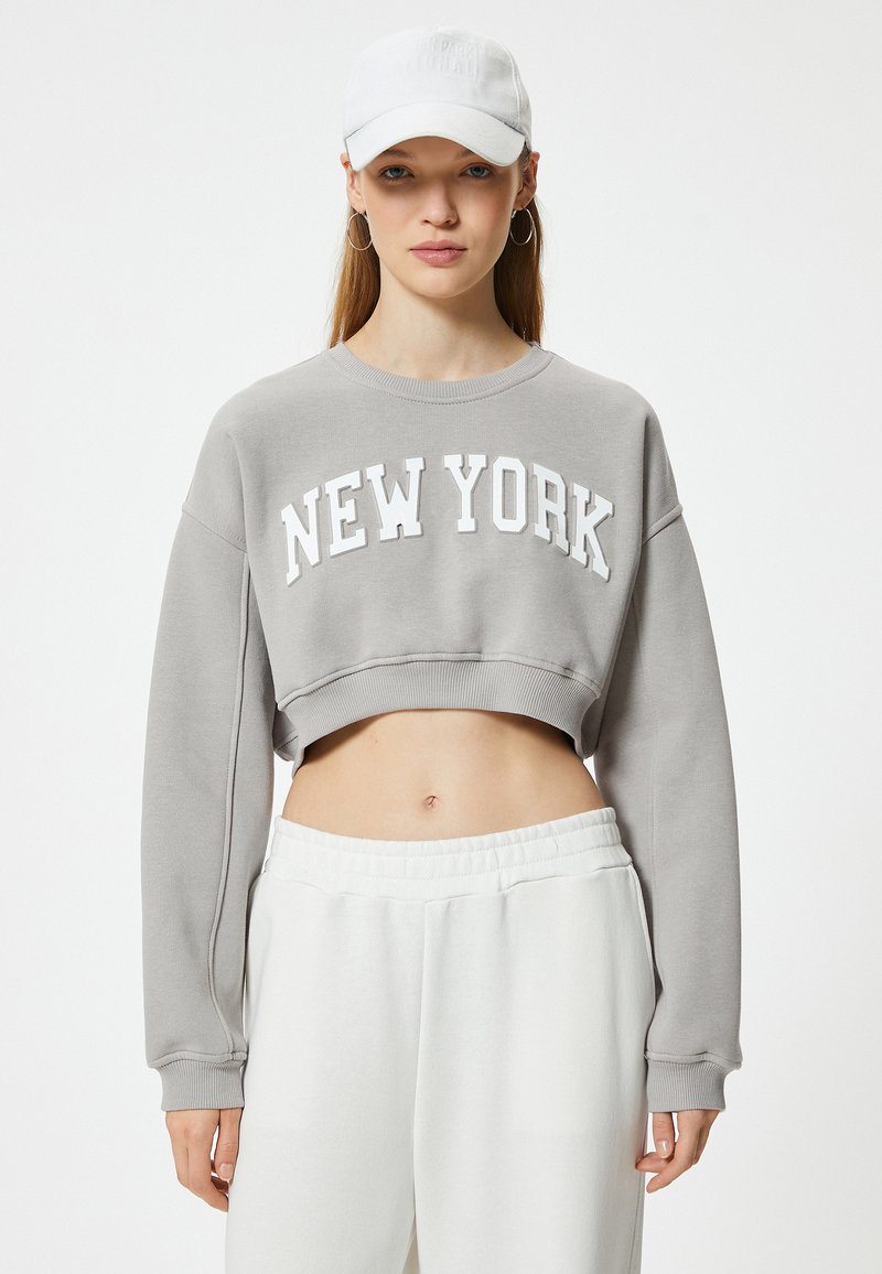 Szary, krótki sweatshirt z napisem "NEW YORK" w białych literach, z długimi rękawami i strukturalnym wykończeniem; połączony z białymi spodniami z elastycznym pasem.