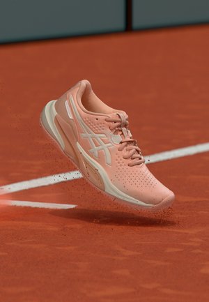 Roze sportschoen in de lucht boven een rood gravel tennisbaan met witte grenslijnen, waarbij kleine graveldeeltjes omhoog worden getrapt.