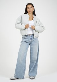 Jasnoszara kurtka bomberka z wysokim kołnierzem, zamkiem błyskawicznym, elastycznymi mankietami, zestawiona z szerokimi niebieskimi dżinsami i szarymi trampkami.