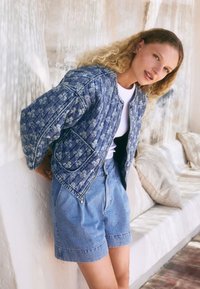 Denim bomberjakke med blomsterprint, ribstrikkede manchetter og lynlåslukning. Kombineret med højtaljede blå denimshorts. Let tekstur.
