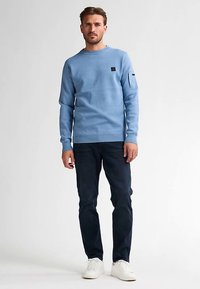 Pull bleu clair en coton, avec un patch noir sur la poitrine, associé à un jean foncé et des baskets blanches. Encolure ronde classique.