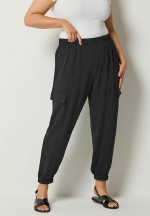 Femme portant un pantalon cargo à taille élastique noire avec poches latérales et des sandales noires, pointant vers la taille sur un fond blanc.