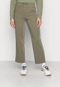 Calças desportivas verde oliva com cintura ajustável, apresentando bolsos laterais e um design de perna reta, combinadas com ténis brancos e beges.