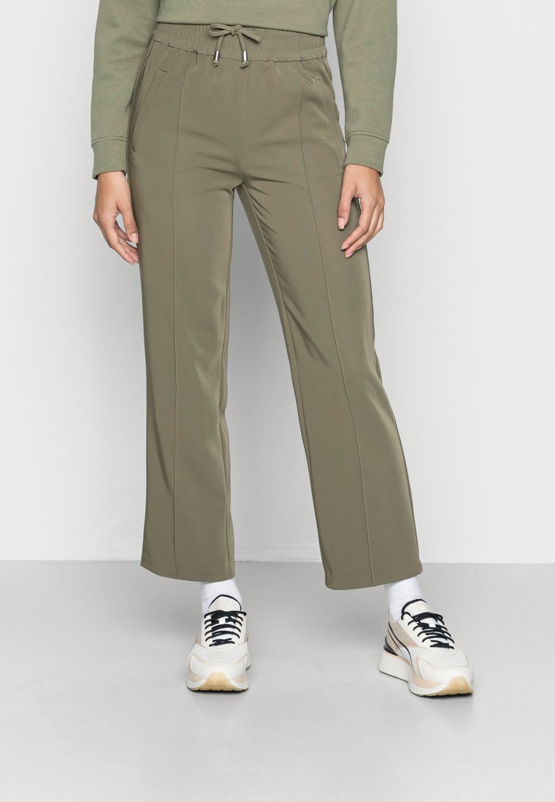 Calças desportivas verde oliva com cintura ajustável, apresentando bolsos laterais e um design de perna reta, combinadas com ténis brancos e beges.