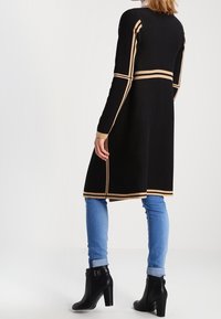 Schwarzer Strickcardigan mit beigen Streifen, mittellang, taillierter Schnitt. Getragen über hellblauen Jeans und schwarzen Stiefeletten mit einer Schnallendetail.