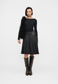 Femme portant un haut noir à manches longues, une jupe plissée en jean foncé avec double boucle, un sac à épaule noir en fourrure et des bottes hautes brun foncé jusqu'aux genoux.