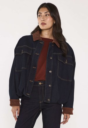 Donna con lunghi capelli scuri indossa una giacca di jeans scura oversize con colletto e polsini in velluto a coste marrone sopra una maglia color ruggine e jeans scuri.