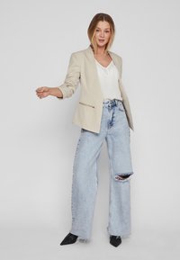 Sacou bej cu rever și buzunar cu fermoar, asortat cu un top alb și blugi evazați de culoare bleu deschis, cu talie înaltă și genunchi distruși. Pantofi negri cu vârf ascuțit.