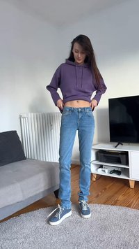 Ljubičasta skraćena majica s kapuljačom, plave traperice i plave tenisice s uzorkom. Okruženje uključuje bež kauč i TV ormar s opremom.