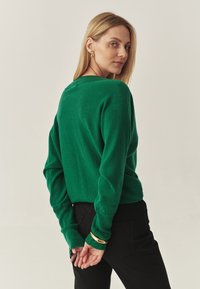 Grüner gestrickter Pullover mit langen Ärmeln und lockerer Passform. Verfügt über gerippte Bündchendetails. Kombiniert mit enganliegenden schwarzen Hosen.