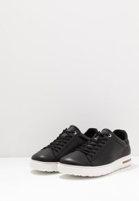 Sneaker in pelle nera con suole bianche, punta rotonda e design con lacci. Presenta un branding sottile e un accento in sughero sul lato.