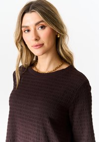 Brown textured top with a crew neckline, featuring a ribbed pattern. Paired with a chunky gold chain necklace. Soft fabric, relaxed fit.