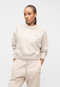 Νεαρή γυναίκα με πλεξούδες, που φοράει ανοιχτό μπεζ φούτερ και αντίστοιχο παντελόνι φόρμας, με τα χέρια στις τσέπες, κοιτάζοντας ελαφρώς προς τα πλάγια.