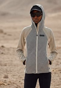 Osoba odjevena u bež prsluk s prugama i patentnim zatvaračem, s rukama u džepovima, crnom kapom i velikim crnim sunčanim naočalama u pješčanom vanjskom okruženju.