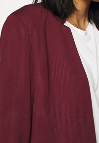 Femme portant un blazer côtelé bordeaux sur un t-shirt à col rond blanc, avec des cheveux bruns atteignant les épaules.