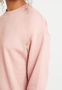 Světl růžová mikina s kulatým výstřihem, volným střihem a hebkou strukturou, zobrazena z boku, aby vynikl design rukávu.