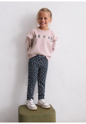 Lichtroze sweatshirt met ruches aan de mouwen en "Marc O'Polo" tekst, gecombineerd met donker patroon leggings en witte sneakers, staande op een groene kruk.