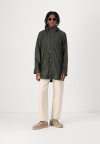 Olijfgroene waterdichte jas met capuchon, drukknoopsluitingen en zijzakken; gecombineerd met lichtbeige broeken en tan schoenen.