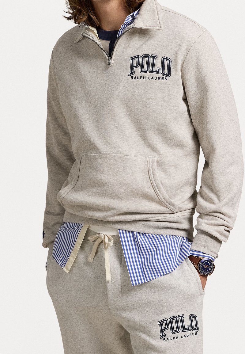 Beige pullover sweatshirt med dragkedja vid kragen, framficka och marinblå "POLO RALPH LAUREN"-logotyp. Kombinerad med gråa sweatpants och randig skjorta under.