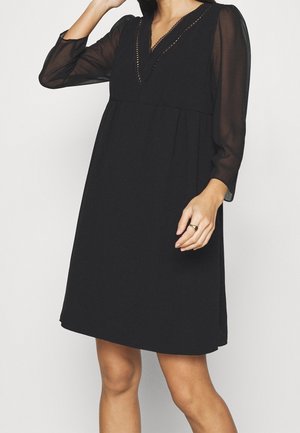 Kvinde iført en knælængde sort kjole med gennemsigtige lange ærmer og en V-udskæring med dekorativ kant, poserer mod en ensfarvet baggrund.