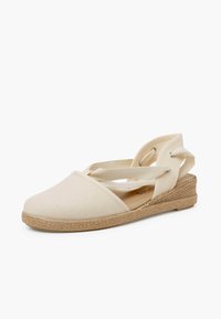 Espadrilla in tela beige con cinturini incrociati e suola in corda juta, caratterizzata da un design piatto e una punta rotonda.