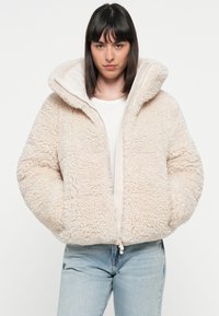 Crème-kleurig fleece jack met een capuchon, gestructureerde stof, getailleerde pasvorm, ritssluiting en zijzakken. Draagt over een wit shirt.